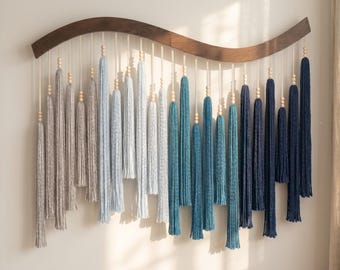 May include: A wall hanging featuring a curved dark wood bar with multiple hanging cotton cords. Each cord is adorned with small wooden beads and ends in a long tassel. The tassels transition in color from light gray to blue and dark navy.