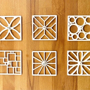 Könnte beinhalten: Sechs weiße quadratische Wandplaketten mit verschiedenen geometrischen Designs. Jede Plakette hat einen kleinen runden Knopf in der Mitte.