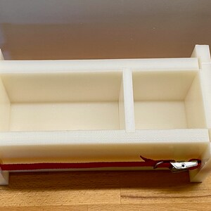 Può includere: Stampo per sapone in plastica bianca con due scomparti e un cinturino rosso con una fibbia argentata.