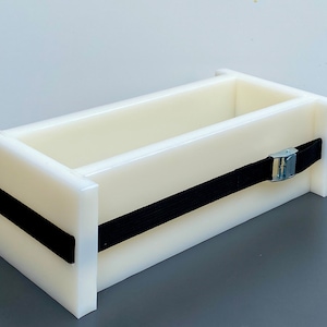 Può includere: Uno stampo per sapone rettangolare bianco sporco con un cinturino nero e una fibbia metallica. Lo stampo è progettato con bordi dritti e un interno cavo, ideale per la creazione di saponi fatti a mano. Il cinturino nero fissa i lati.