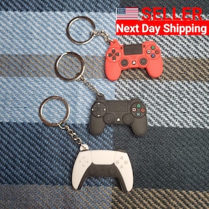 May include: Three keychains featuring different colored PlayStation controller designs. The keychains are red, black, and white. The keychains are on a blue and white plaid background.