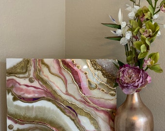 Könnte beinhalten: Ein quadratisches Leinwandbild mit abstrakten, achatähnlichen Mustern in Rosa-, Weiß- und Goldtönen. Das Bild wird auf einem Tisch neben einer Vase mit rosa Blumen und grünen Blättern ausgestellt.