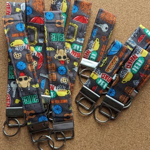 May include: A set of six colorful keychains featuring a Friends theme. The keychains are made of fabric and feature various iconic images from the show, including the Central Perk logo, the iconic purple door, and the phrase "How You Doin'?" The keychains are attached to silver key rings.