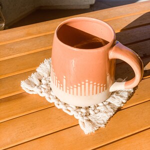 May include: A pink ceramic mug with a white geometric design sits on a white crocheted coaster with fringe. The mug and coaster are on a wooden surface.