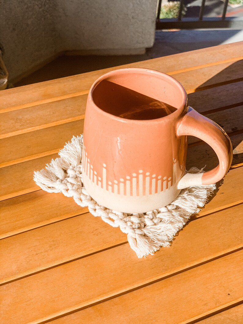 May include: A pink ceramic mug with a white geometric design sits on a white crocheted coaster with fringe. The mug and coaster are on a wooden surface.