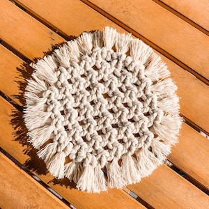 May include: A round, cream-colored crocheted coaster with a fringe border. The coaster is made of cotton cord and has a woven pattern.