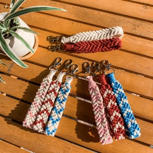 May include: A collection of macrame keychains in various colors, including cream, red, pink, and blue. Each keychain features a woven design and a silver-colored clasp. The keychains are arranged on a wooden surface.