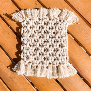 May include: A white crocheted coaster with fringe on all sides. The coaster is on a wooden surface.