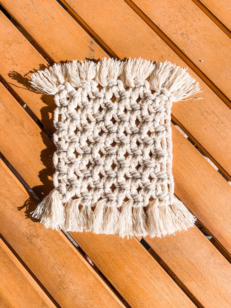 May include: A white crocheted coaster with fringe on all sides. The coaster is on a wooden surface.