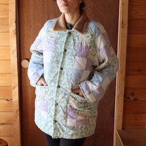 May include: A quilted jacket with a patchwork design in shades of blue, lavender, and white. The jacket features a brown collar, wooden buttons, and two front pockets. The floral pattern adds a vintage touch to this outerwear.