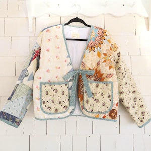 May include: A patchwork quilted jacket with a cream-coloured base and floral and geometric patterns in blue, brown, and orange. It has two front pockets and a light blue tie closure. The jacket is hanging on a black hanger.