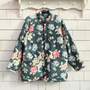 May include: A dark green quilted jacket with a floral pattern of pink, white, and yellow flowers. The jacket has a high collar, button closure, and two front pockets. The jacket is hanging on a metal hanger.