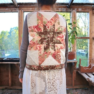 May include: A sleeveless waistcoat featuring a large, central star-shaped quilt pattern on the back. The design incorporates floral fabric in shades of pink, red, and brown, contrasted with off-white textured panels and a dark brown floral border trim.