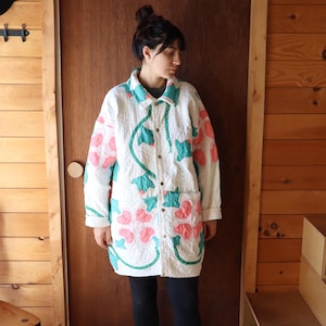 May include: White quilted jacket with pink and teal floral appliqués. The jacket has a button-down front, two pockets, and a collar. The floral design is outlined with green stitching. This long coat has a unique, artistic look.