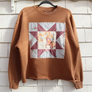 May include: A brown sweatshirt with a patchwork design. The patchwork features a star pattern with a butterfly and floral print. The sweatshirt has a crew neck and long sleeves.