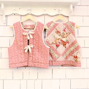 May include: Two quilted vests hanging on wooden hangers. The front vest is pink with a small white floral pattern and two cream-colored ribbon ties. The back vest features a patchwork star design with floral fabrics in shades of pink, green, and cream.