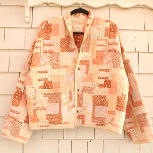 Peut inclure: Veste matelassée à patchwork aux tons pêche, crème et marron. Elle présente un col, deux poches avant et des boutons. Le design est composé de divers carrés de tissu, pour un style vintage et artisanal.