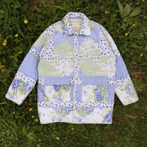 May include: A long-sleeve quilted jacket featuring a patchwork design of blue and white floral patterns, light green and white stripes, and blue and white gingham checks. The jacket has a collared neckline, a button-down front, and two pockets.