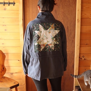 May include: A dark grey jacket with a star-shaped patch on the back. The patch features a floral and butterfly design on a white background, framed by a dark green floral pattern. The jacket has a drawstring waist and a high collar.