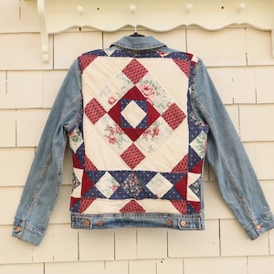 May include: A blue denim jacket featuring a quilted back panel. The quilt design consists of cream, burgundy, and navy blue fabric patches with floral patterns arranged in a geometric block layout. The jacket has long sleeves and a collar.