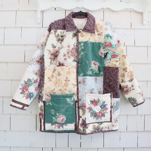 May include: A patchwork jacket with a floral pattern in shades of cream, green, and purple. The jacket features a button-up front, two pockets, and a quilted design. The sleeves and collar are trimmed with a dark brown fabric.