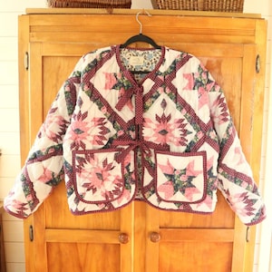 Peut inclure: Une veste matelassée au motif patchwork avec des imprimés floraux roses et rouge foncé sur fond blanc. La veste présente une encolure ronde, des manches longues, deux poches avant et deux attaches en tissu sur le devant.
