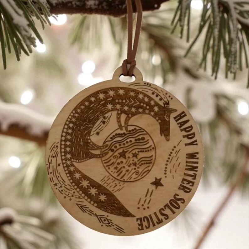 May include: A round wooden ornament with a fox design and the words "Happy Winter Solstice" engraved around the edge. The ornament is hanging from a brown leather cord. The background is blurred with Christmas lights.