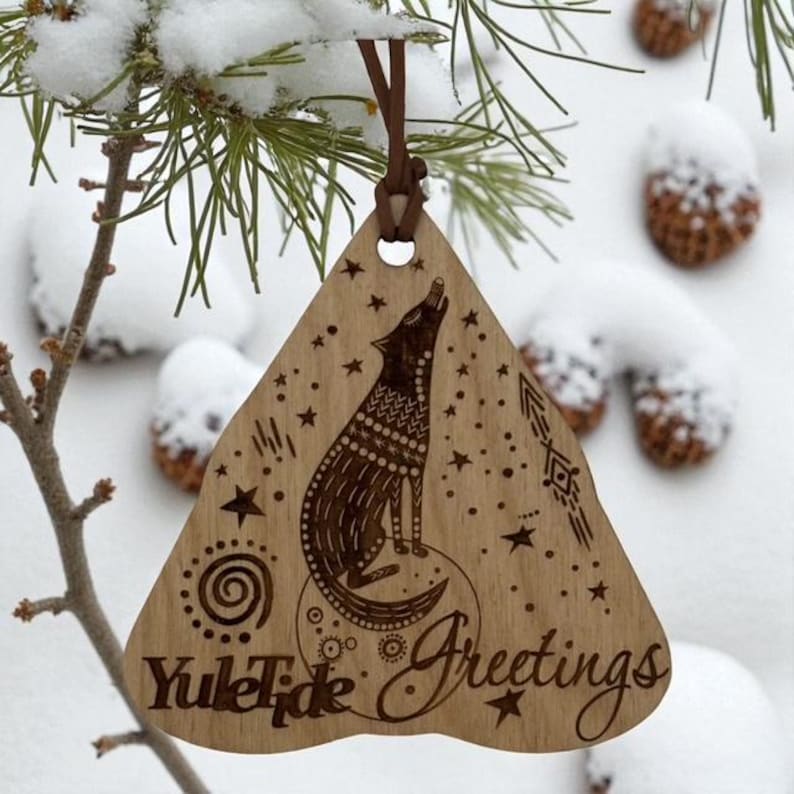 May include: Wooden Christmas ornament with a howling wolf design, stars, and the words "Yule Tide Greetings." The ornament is triangular, with a leather cord for hanging. The background shows snow-covered pine needles and acorns.