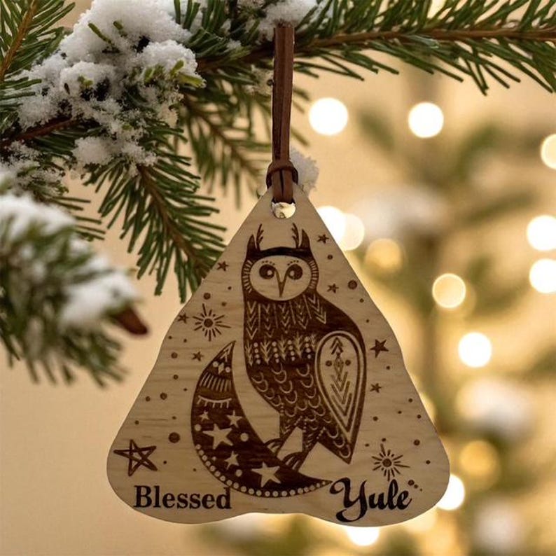 May include: Wooden Christmas ornament with an owl design. The ornament is triangular with a brown leather strap. The owl is etched into the wood, along with stars, a crescent moon, and the words "Blessed Yule."