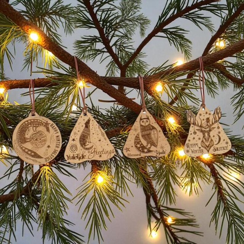May include: Wooden Christmas ornaments with engraved designs of a deer, owl, fox, and a circular design. Each ornament has a different holiday-themed message, such as "Happy Winter Solstice" and "Yuletide Greetings". The ornaments are hanging on a pine branch with fairy lights.