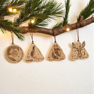 May include: A collection of four wooden Christmas ornaments hanging from a pine branch. The ornaments feature engraved designs of a crocodile, fox, owl, and deer, with festive text. Each ornament is suspended by a brown string.