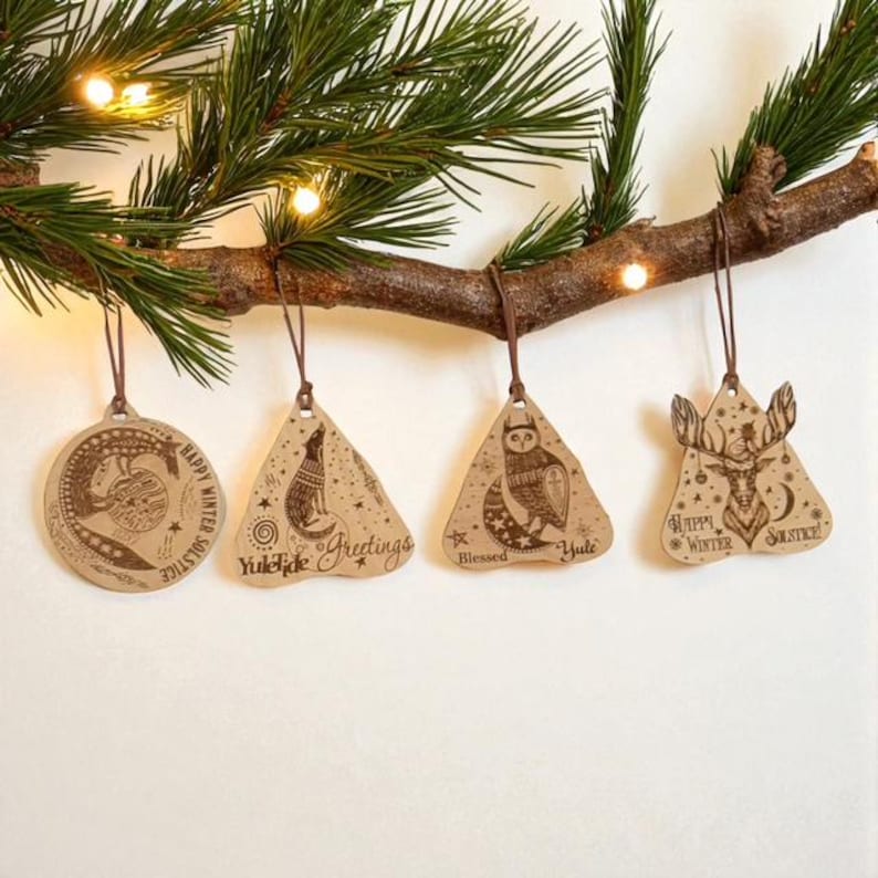 May include: A collection of four wooden Christmas ornaments hanging from a pine branch. The ornaments feature engraved designs of a crocodile, fox, owl, and deer, with festive text. Each ornament is suspended by a brown string.