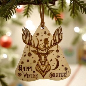 May include: A wooden Christmas ornament in a triangular shape, featuring a detailed engraving of a deer with antlers wearing a hat. The ornament has the words "Happy Winter Solstice!" and is decorated with stars and snowflakes.