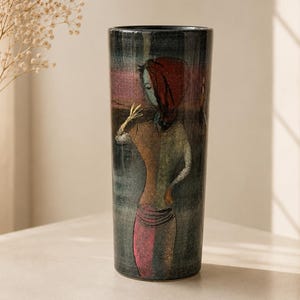 Può includere: Un vaso alto e cilindrico con un'illustrazione dipinta di una donna con un copricapo e un vestito rossi. Il vaso ha uno sfondo scuro e strutturato con sfumature di rosa e verde. L'opera d'arte ricorda un dipinto vintage.