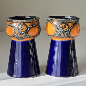 Puede incluir: Dos candelabros de cerámica. La base es azul oscuro, ensanchándose desde una base estrecha. La parte superior es naranja con un diseño negro en espiral. Los bordes son negros. Los candelabros están sobre una superficie blanca.