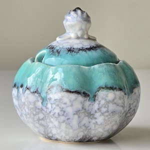 Könnte beinhalten: Ein Keramiktopf mit Deckel. Der Topf ist rund mit einem türkisfarbenen Rand und einem gesprenkelten grau-weißen Körper. Der Deckel hat einen kleinen, dekorativen Knopf. Der Topf wird wahrscheinlich zur Aufbewahrung kleiner Gegenstände verwendet.