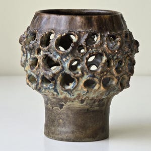 May include: A brown ceramic vase with a textured, porous design around the middle. The vase has a wide opening and a short, cylindrical base. The color palette includes shades of brown, green, and blue.