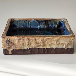 May include: A square ceramic dish with a textured, layered design. The top interior is a deep blue, transitioning to a tan and brown exterior. The bottom section is a dark brown. The dish is likely handmade.