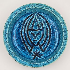 May include: A round, blue ceramic bowl with a textured surface. The bowl features a central black design of a stylized face and star. The bowl's rim has a lighter blue color.