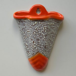 Könnte beinhalten: Eine weiße Keramik-Wandvase mit orangefarbenem Rand und einer strukturierten Oberfläche. Die Vase hat oben ein Loch zum Aufhängen.