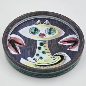 Peut inclure: Un plat rond en céramique au design fantaisiste. Le plat présente un fond bleu foncé avec un visage stylisé au centre, des yeux verts et des accents rouges. Le bord est marron foncé et vert.