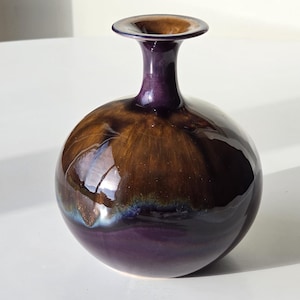 Puede incluir: Un pequeño jarrón de cerámica redondo con cuello estrecho y borde acampanado. La superficie presenta un esmalte brillante con una mezcla de tonos violeta intenso y marrón, creando un acabado suave y reflectante en el cuerpo esférico.