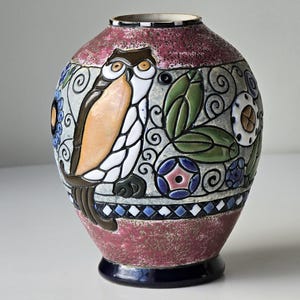 Op de afbeelding: Een decoratieve keramische vaas met een afgeronde body en een donkerblauwe basis. De vaas heeft een vogelontwerp met oranje, witte en bruine details, omgeven door bloemen en geometrische patronen in blauw, groen en wit. De bovenste rand is wit.