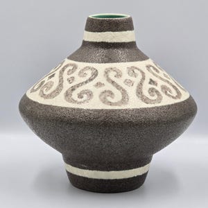 May include: A decorative ceramic vase with a unique shape and design. The vase features a dark brown base and a cream-colored upper section with a repeating scroll pattern. White stripes encircle the neck and base. The interior is green.