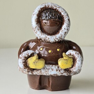 May include: A brown ceramic figurine of a person wearing a hooded parka with a white fur trim. The figure has yellow mittens and is posed with hands clasped. The parka has gold-colored buttons.