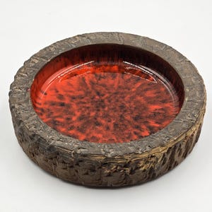 Può includere: Un posacenere rotondo in ceramica con un esterno marrone scuro strutturato e un interno rosso vibrante. L'interno presenta un motivo maculato, che aggiunge interesse visivo. Un oggetto decorativo.