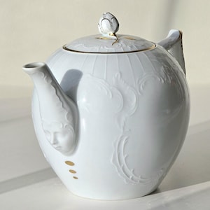 Puede incluir: Tetera de porcelana blanca con un diseño floral dorado y un pomo dorado en forma de capullo en la tapa. La tetera tiene una cara esculpida en el pico.