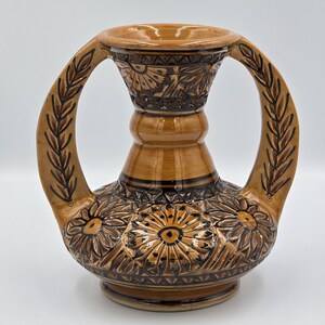 Può includere: Un vaso in ceramica marrone con due manici ad arco e un'ampia apertura. Il vaso presenta intricati motivi floreali e di foglie intagliati sulla superficie. I manici sono adornati con disegni di foglie.