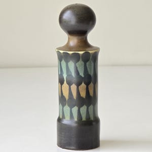 Könnte beinhalten: Dunkelbrauner Keramik-Flaschenverschluss mit rundem Kopf und zylindrischem Körper. Der Körper weist ein Muster aus teal, gold und schwarzen Formen auf. Ein dekorativer Artikel.
