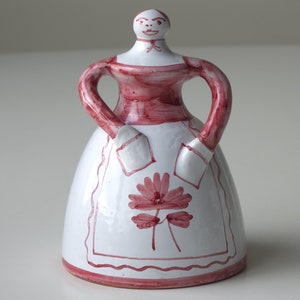 Puede incluir: Una figurita de cerámica de una mujer con un vestido blanco con un diseño de flores rojas y un corpiño rojo. La figurita tiene una cara pintada simple y está de pie sobre una superficie blanca.